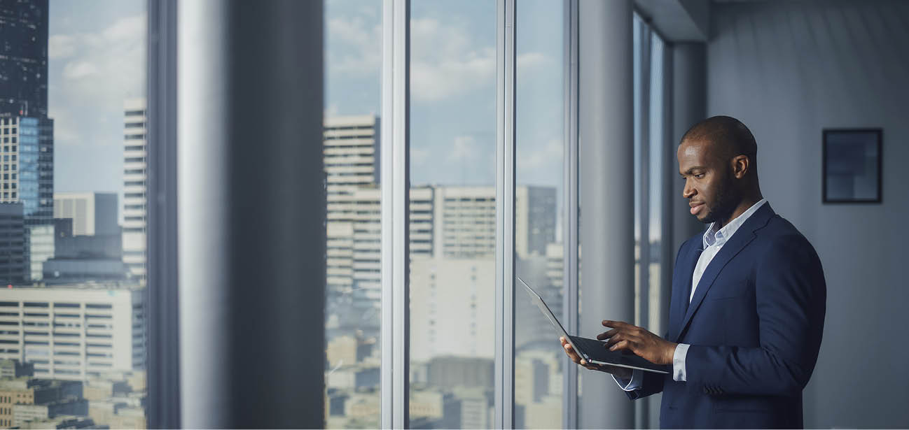 Thoughtful Black Businessman in a Tailored Suit Using Laptop while Standing in Office Near Window on Big City. Successful Corporate Top Manager Doing Data Analysis for e-Commerce Startup