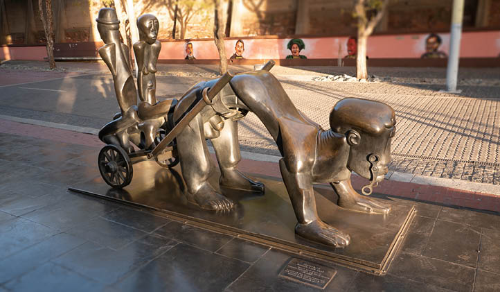 South African artist, died in exile in New York.   This large sculpture, which is installed at the entrance to the Constitutional Court, was initially a much smaller clay artwork made in 1987 by Feni. In 2003 it was enlarged and cast in bronze by the Tallix Art Foundry in Brooklyn, New York and shipped to South Africa. The work depicts the brutality of the master-slave relationship: a large yoked figure draws a cart upon which two other figures sit. In fact their seat is made of another human figure. Drawing on the title of the work, History, it suggests the brutal treatment of the black body during apartheid and the broader harnessing of black labour in the colonial enterprise.