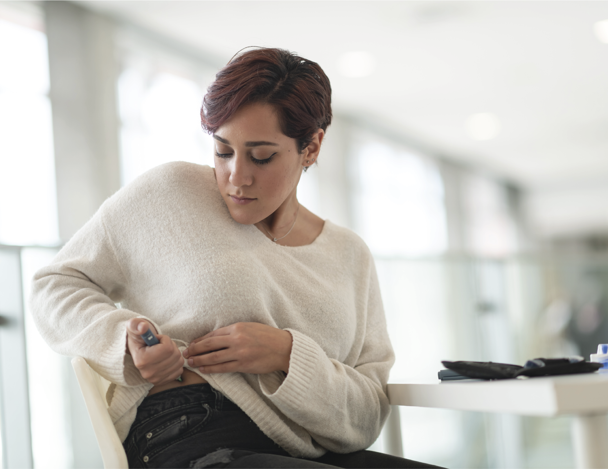 A woman of Middle Eastern ethnicity is seated at a table. She is taking a break from work and is in her workplace's lobby. The woman is diabetic and is using an insulin pen near her hip.