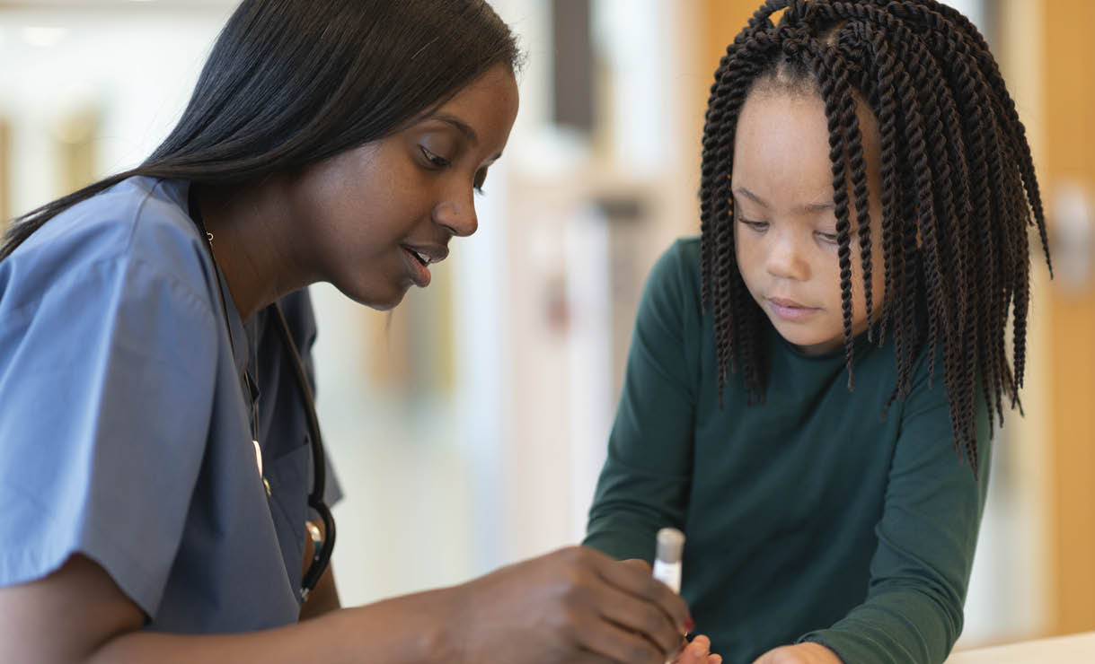 An African American medical professional meets with a patient. The patient is a young diabetic girl. The two are seated at a table together. The doctor happily explains how to use an insulin pen. The girl is watching intently.