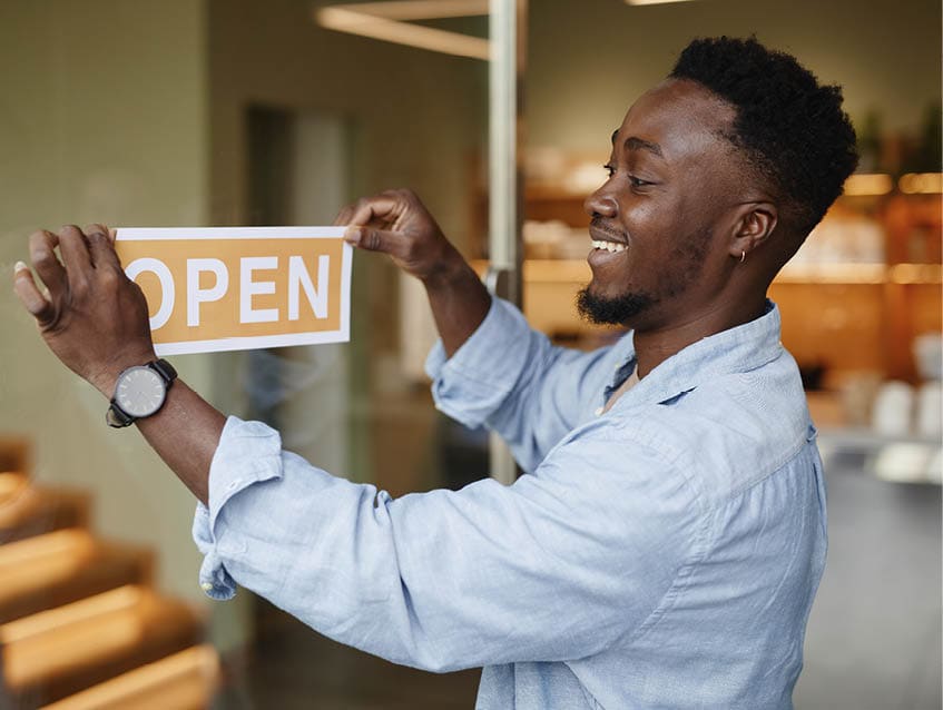 Joyful young African American man opening modern cafe in morning sticking sign on glass door