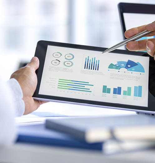 Two business men meeting and looking at financial data, charts and graphs on a digital tablet and a laptop computer. There are documents and paperwork on the table. One man is pointing at the screen